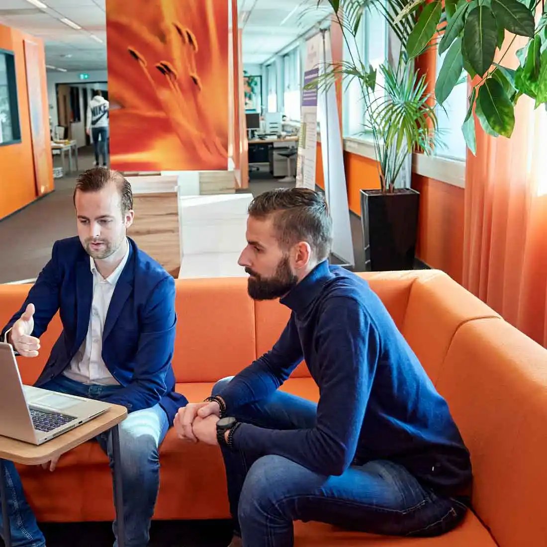 Twee mannen zitten op oranje bank naar een laptopscherm te kijken.